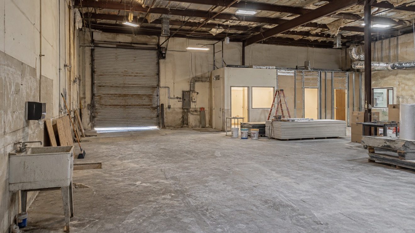 An industrial suite before work, with torn ceiling insulation, exposed conduit, a stained concrete slab, and a corroded roll-up dock door.